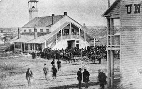 Black and white photograph of a crowd gathered in front of a large white wooden building, quite imposing for the period. The second floor has a balcony accessible via external staircases.