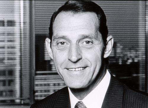 Photographie en noir et blanc d’un homme d’âge moyen souriant, en costume-cravate. L’homme est devant une fenêtre, avec vue sur des édifices en hauteur.