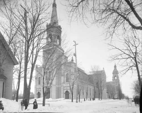 Photographie en noir et blanc d’une église vue en hiver. Une maison, des arbres et quatre personnes font partie du paysage. Une partie de la photographie est masquée, à cause de l’objectif photographique.