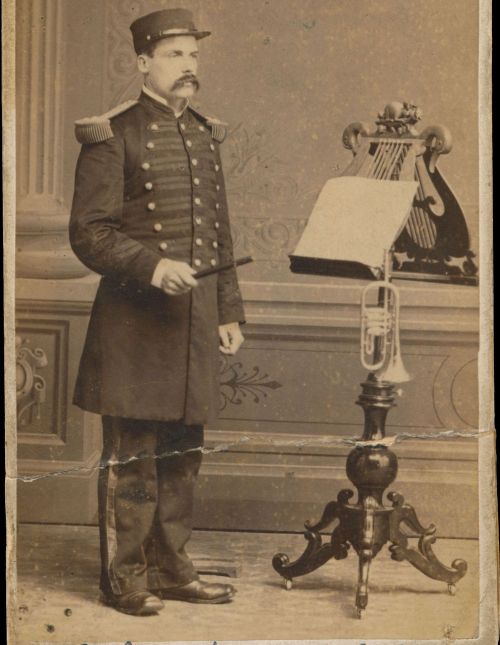 Photographie sépia d’un homme d’âge moyen. Il porte un uniforme et est debout devant un lutrin richement sculpté où reposent des partitions de musique et une trompette. Le nom et le titre du sujet sont écrits à la main au bas de la photo.