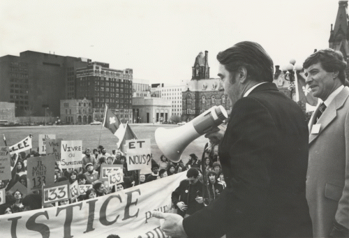 Photographie en noir et blanc de deux hommes d’âge moyen, dont l'un tient un porte-voix. Il s'adresse à un groupe de manifestants, qui brandissent des pancartes, des drapeaux franco-ontariens et une large banderolle. À l'arrière plan, l'édifice de l'Ouest et la rue Wellington.