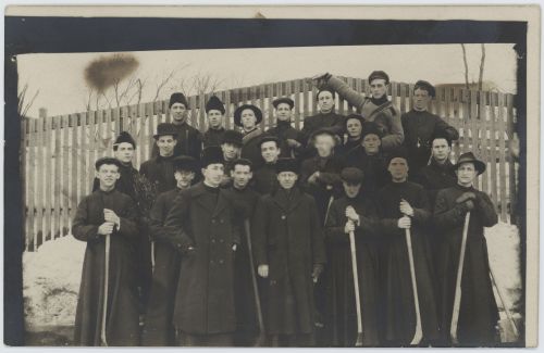 Black and white photograph, taken in winter, of a group of about fifteen young men, accompanied by some adults. They are arranged in three rows, in front of a wooden fence. The youngest wear the cassock and hold hockey sticks. Adults wear long winter coats and fur hats. One of them stands in front of the group, the other in the center of the photo, the other two on the sides.