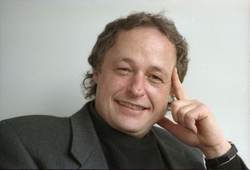 Photographie en couleur d’un homme souriant d’âge moyen, en costume foncé. Il est assis, son index gauche est appuyé contre sa tempe et son pouce gauche, contre sa mâchoire.