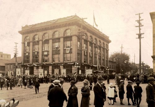 Photographie en noir et blanc d'une foule massée de chaque côté d'une rue. Hommes, femmes et enfants sont habillés élégamment. À l'arrière, un bâtiment de quatre étages, richement décoré de banderolles et drapeaux. Plusieurs étendards à l'effigie de la Vierge et autres saints patrons.