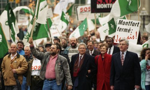 Photographie en couleur d’une manifestation, en pleine rue. Un manifestant porte une pancarte qui se lit : «Montfort fermé : Jamais ! ». La foule agite des drapeaux franco-ontariens.