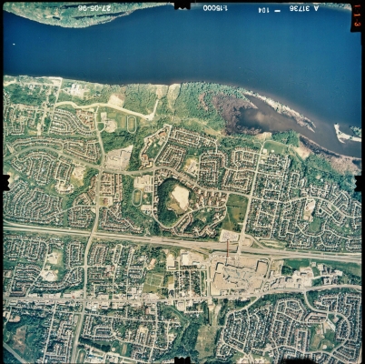 Photographie aérienne en couleur d’un paysage de banlieue où se trouve, au nord (au haut de l'image), une rivière. De nombreuses habitations sont regroupées en lotissements. Une autoroute sépare la zone en deux. On distingue un grand centre commercial au centre de la photo.