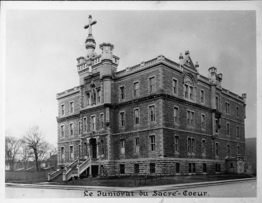 Black and white photograph with text printed in French. A three-storey brick building, with many windows, surmounted by an imposing cross. A staircase leads to the main entrance.