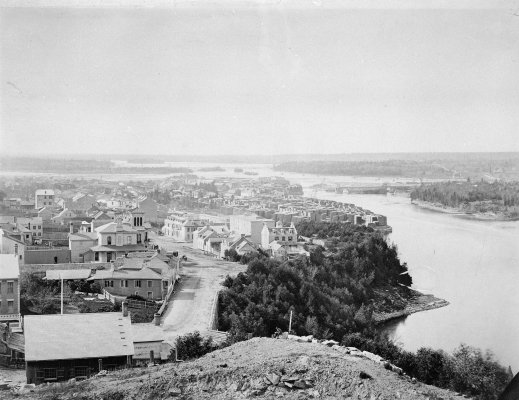 Photographie en noir et blanc d’un paysage urbain, vu de haut. Une imposante rivière le borde à droite, traversée par un pont qui mène à une zone boisée. Au loin, à perte de vue, la forêt.
