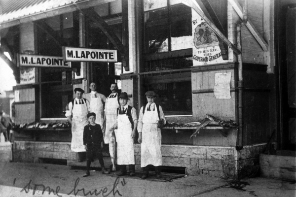 Photographie en noir et blanc de cinq hommes en tablier blanc et d’un garçon devant une poissonnerie. Des enseignes indiquent la raison sociale de l'entreprise ainsi que sa distinction en tant que fournisseur du Gouverneur général. “Some bunch” est écrit à la main en anglais, au bas de la photo.