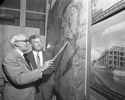 Photographie en noir et blanc d’un homme d’un certain âge et d’un homme d’âge mûr, tous deux en costume et cravate. Les deux hommes sourient en regardant une grande carte aérienne très détaillée, accrochée au mur. Le plus âgé des deux hommes se sert d’une règle pour indiquer un endroit précis sur la carte.