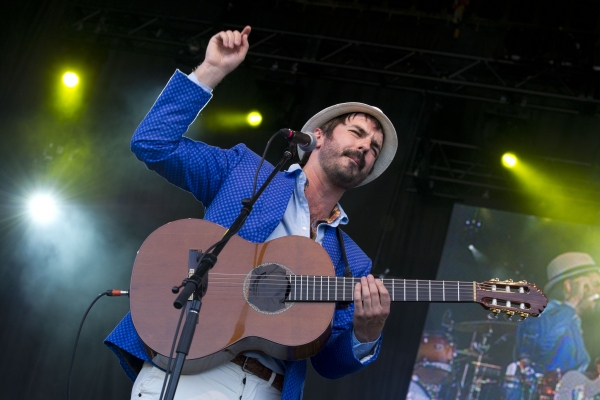 Photographie en couleur d’un homme d’âge moyen, sur une scène. Il porte une barbe courte, noire, un chapeau, un veston bleu royal et un pantalon blanc.  Il joue de la guitare acoustique et chante devant un microphone. Il est photographié en contre-plongée et son image se reflète sur une surface, à sa gauche.