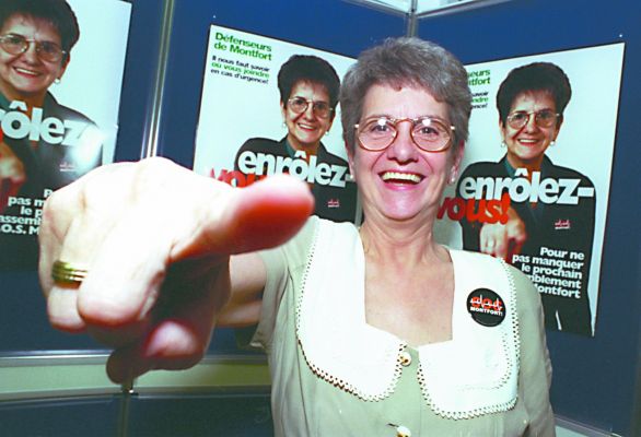 Colour photograph of a mature woman, smiling and mimicking the gesture she herself is making on the posters behind her. On the poster she wears an S.O.S. Montfort button and points her finger at the camera. Superimposed on her chest, the slogan: "Enrôlez-vous !'