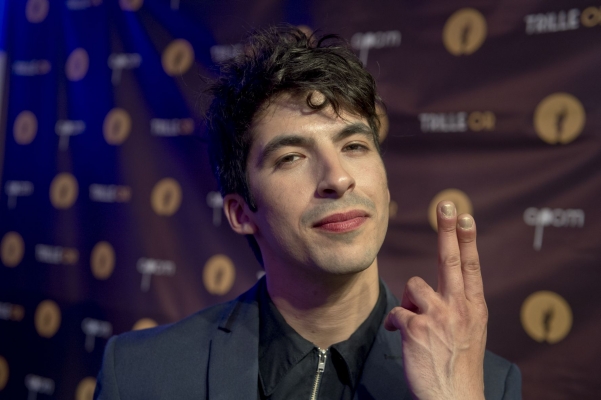 Colour photograph of a mature man with disheveled black hair, wearing a gray jacket and a black shirt. He holds up his left hand, pointing up with the index and middle fingers. The blue-lit wall in the background is covered in brown cloth with golden circles and words in white letters.