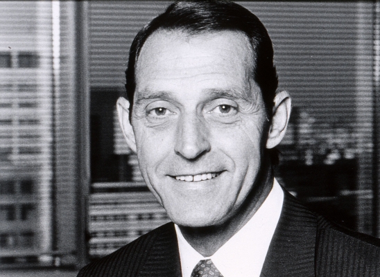 Photographie en noir et blanc d’un homme d’âge moyen souriant, en costume-cravate. L’homme est devant une fenêtre, avec vue sur des édifices en hauteur.