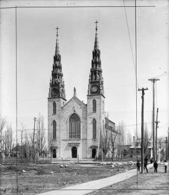 Photographie en noir et blanc d’une grande église en pierre blanche à trois étages avec, au centre de la devanture, un large vitrail. L’église est ornée de deux clochers. L’espace devant l’église reste vacant. Quelques petites demeures, ainsi que des arbres et des fils électriques se trouvent de chaque côté de l’église.