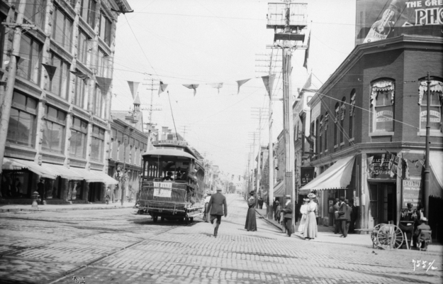 Photographie en noir et blanc d’un tramway qui circule sur un large boulevard commercial décoré de banderoles. Des gens marchent le long des trottoirs et montent dans le tramway.