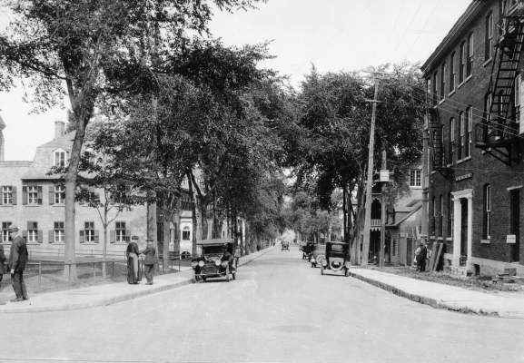 Photographie en noir et blanc d’une rue passante bordée d’arbres, avec de nombreuses automobiles et des piétons. Des résidences se trouvent d’un côté de la rue et un édifice institutionnel de l’autre, devant lequel des hommes, dont un religieux, discutent.