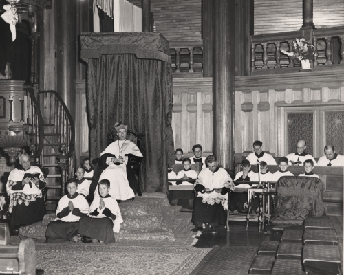 Photographie en noir et blanc d’un religieux d’âge mûr, portant la barrette, assis dans un fauteuil surélevé à côté de la chaire. Il est entouré de cinq autres religieux et de jeunes servants de messe, tous habillés en soutane et en surplis.