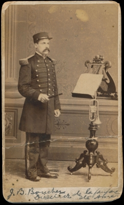 Photographie sépia d’un homme d’âge moyen. Il porte un uniforme et est debout devant un lutrin richement sculpté où reposent des partitions de musique et une trompette. Le nom et le titre du sujet sont écrits à la main au bas de la photo.