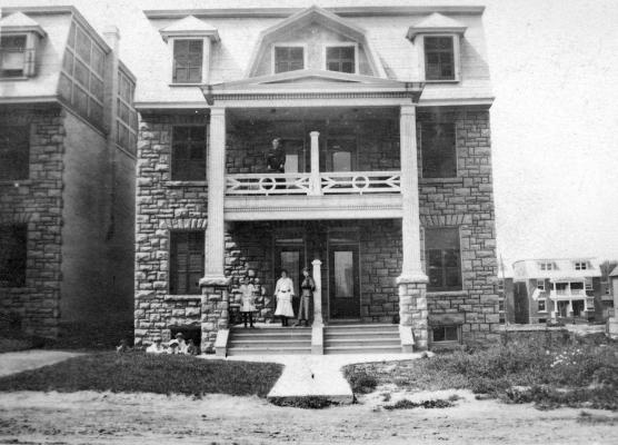 Photographie en noir et blanc. Femmes et enfants devant deux maisons attenantes en pierre à trois étages, avec balcons.