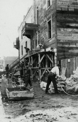 Photographie monochrome sépia de six hommes construisant une maison en bois à trois étages. Le site semble rudimentaire et les travailleurs, peu préoccupés par la sécurité.