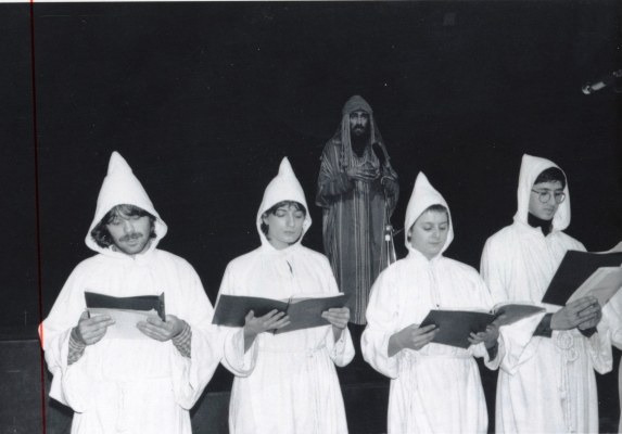 Photographie en noir et blanc de deux jeunes femmes et deux jeunes hommes, tenant des cahiers ouverts. Ils portent des tuniques, avec des capuchons. Debout derrière eux, un homme barbu en tunique rayée multicolore avec un capuchon , devant un microphone.