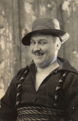 Photographie portrait en noir et blanc d’un homme d’âge mûr souriant. Il arbore la moustache. Il est vêtu d’une chemise épaisse, porte un chapeau et une ceinture fléchée.