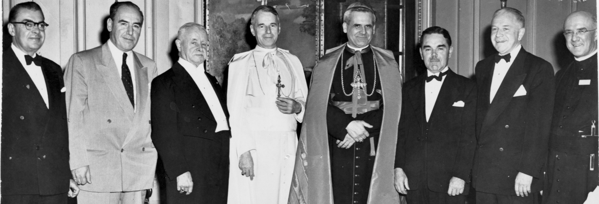Photographie en noir et blanc d’un groupe d’hommes, d’âges divers, en habits de soirée. Le groupe inclut trois religieux portant les vêtements liturgiques d’un évêque pour le premier, ceux d’un archevêque pour le second et la soutane pour le troisième.