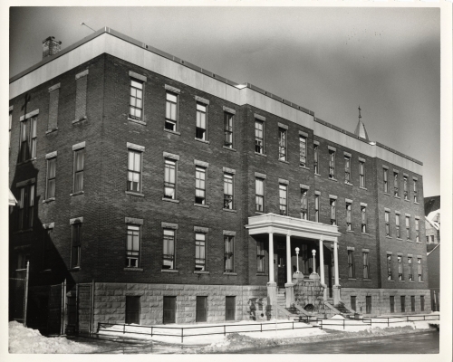 Photographie en noir et blanc d’un édifice en brique à trois étages, percé de nombreuses fenêtres. Deux escaliers séparés mènent à l’entrée principale.