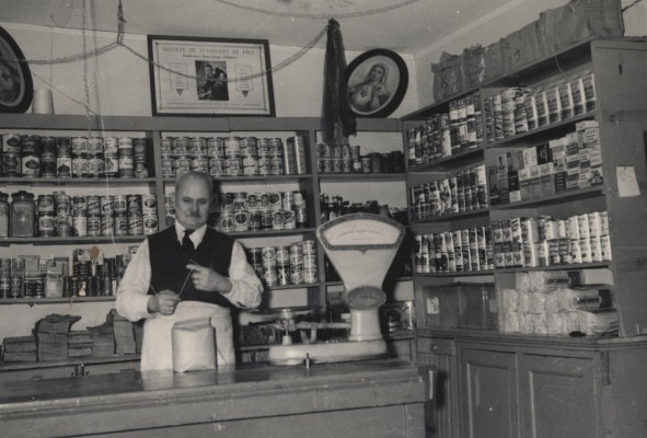 Photographie en noir et blanc d’un homme d’un certain âge, préparant un sac au comptoir d’un magasin. Derrière lui, des étagères remplies de boîtes de conserves et d’autres produits de base.