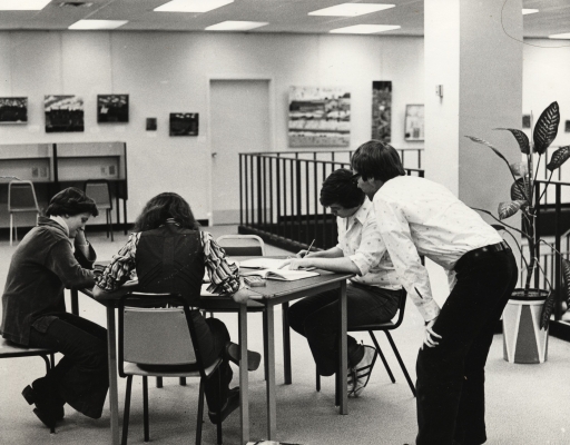 Photographie en noir et blanc d’un groupe de deux jeunes femmes et d’un jeune homme, assis autour d’une table dans une salle d’étude. Un adulte, vu de dos, est penché et observe leur travail.