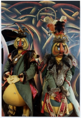 Photographie en couleur de deux personnages déguisés, lors d’une représentation théâtrale. Les comédiens portent des costumes et des masques rembourrés. Ils ont de gros nez orange, de gros yeux et de longs cils. Ils portent des chapeaux flamboyants ainsi que des vêtements garnis de fourrure. À l’arrière-plan, un décor peint avec des motifs de différentes couleurs.