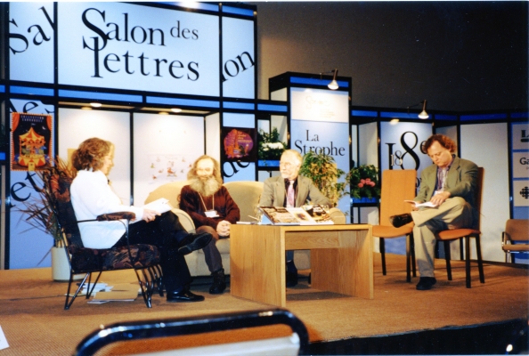 Photographie en couleur d’un groupe de trois hommes et d’une femme, sur une scène. Ils sont assis autour d’une table basse, sur laquelle sont déposés des livres. Le premier se démarque par sa longue barbe rousse. Derrière eux, un décor composé de sections où sont inscrites différentes mentions, dont « Salon des lettres ».