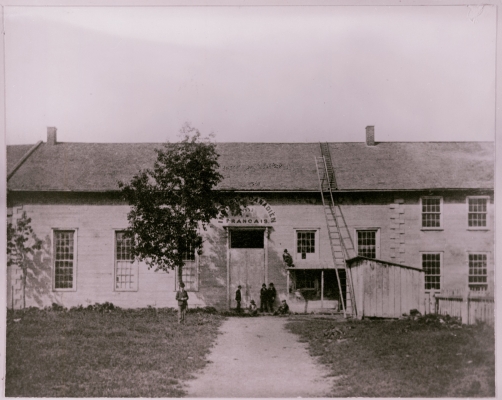 Photographie en noir et blanc d’un long édifice à deux étages en pierre, orné de fenêtres. Au dessus de l’entrée principale, une enseigne de l’Institut canadien-français. Sept hommes se tiennent près de l’entrée principale, dont un juché sur le toit d’un portique.  Une longue échelle rejoint le toit.