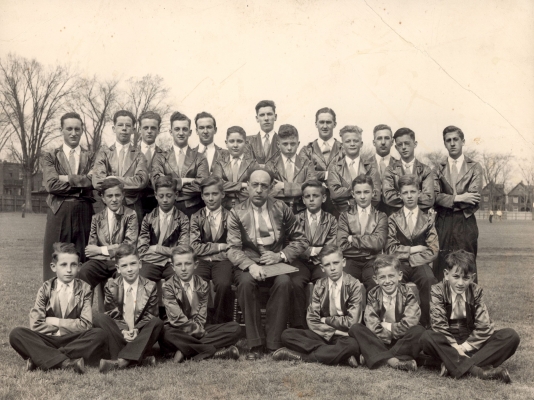 Photographie en noir et blanc d’un groupe de jeunes gens, prise dans un champ. Certains sont assis sur le sol, d’autres sur des chaises, les derniers sont debout à l’arrière.  Un homme d’âge mûr tenant un cartable est assis au centre du groupe. Tous portent un pantalon noir, un veston gris luisant et une cravate.