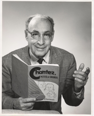 Photographie en noir et blanc, prise en studio, d’un homme d’âge mûr souriant, souffrant de calvitie. L’homme porte des lunettes, un costume à carreaux et une cravate noire. Il tient dans sa main droite un livre, intitulé Chantez, petits et grands.
