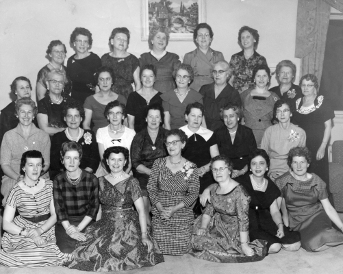Black and white photograph, taken in a living room, of some thirty adult women. They are positioned in four rows, with the first two sitting, and the others standing. Most of the women are middle-aged. All wear elegant dresses and smile at the camera.