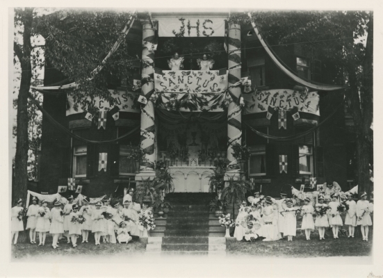 Photographie en noir et blanc d’un reposoir, à l’avant d’une maison de trois étages, richement décorée de bannières et de banderoles. Sur un des balcons, deux statues d’anges. Au pied du reposoir, une trentaine de jeunes enfants habillés de blanc, portant des couronnes et des ailes et tenant des bouquets de fleurs.
