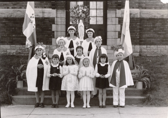 Photographie en noir et blanc d’un groupe d’enfants, d’âges divers. Ils sont debout devant ou sur les marches d’un escalier menant à la porte d’entrée d’un édifice en brique. Ils portent un uniforme scolaire, une robe de communion ou encore un costume spécial avec cape et chapeau assortis.