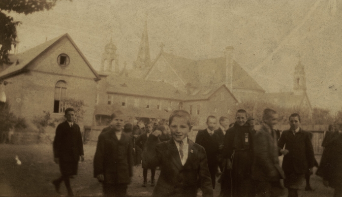 Photographie en sépia d’un groupe d’une vingtaine de jeunes garçons souriants, en costumes et cravates, devant des bâtiments religieux. À l’arrière-plan, des moines discutent avec certains d’entre eux.