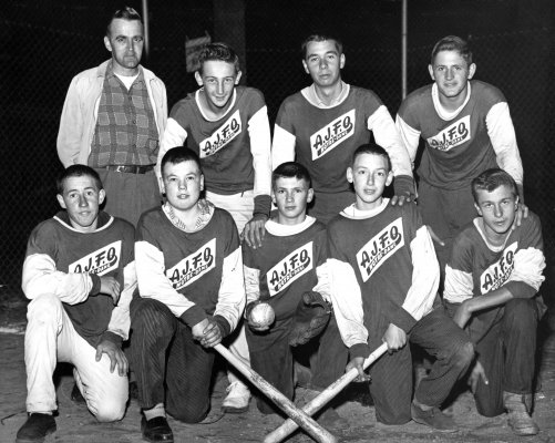 Black and white photograph of a group of eight teens accompanied by a middle-aged man. Five young men are kneeling, with the other three standing behind them. All boys wear “A.J.F.O. Notre-Dame“ jerseys, and three in the front hold baseball equipment.