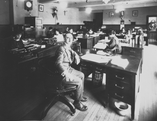 Photographie en noir et blanc d’hommes et de femmes travaillant  à leurs bureaux. À l’avant-plan, un homme d’âge mûr en costume regarde la caméra. À sa gauche, une jeune femme à l’air sérieux, derrière son bureau.