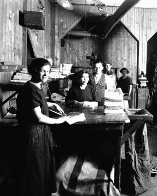 Photographie en noir et blanc de trois jeunes femmes et de deux jeunes hommes debout à des comptoirs. Ils sont entourés de piles de journaux.