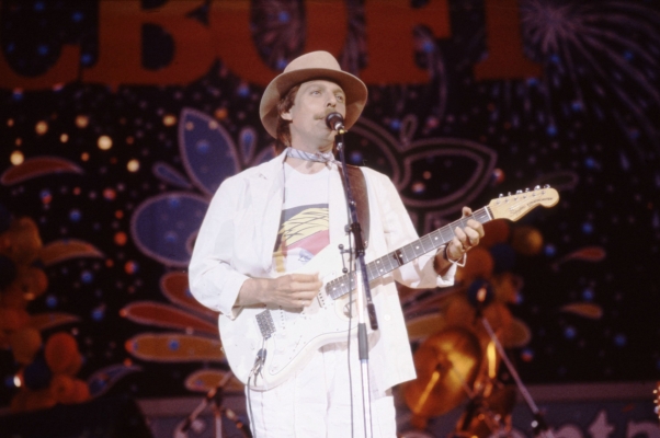 Photographie en couleur d’un homme d’âge moyen moustachu, sur une scène. Il porte un chapeau, un foulard au cou, un costume blanc et un tee-shirt avec un motif coloré. Il joue de la guitare électrique et chante devant un microphone. En arrière-plan, des motifs géométriques sur fond coloré et l’inscription « CBOF-FM ».