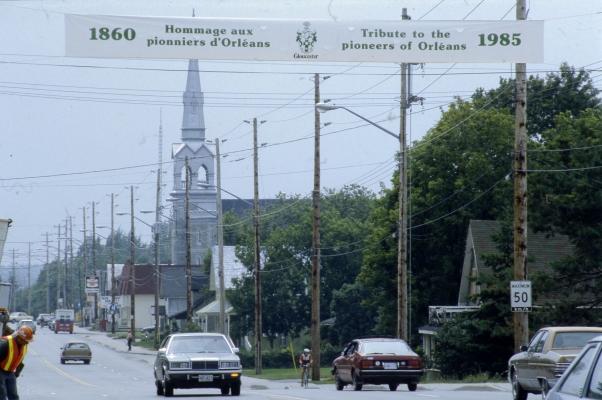 Photographie en couleur d’une rue passante où se côtoient espaces commerciaux, espaces résidentiels ainsi qu’une église. Sur une bannière installée au-dessus de la rue, on lit : « 1860 Hommage aux pionniers d’Orléans/Tribute to the pioneers of Orléans 1985 ».