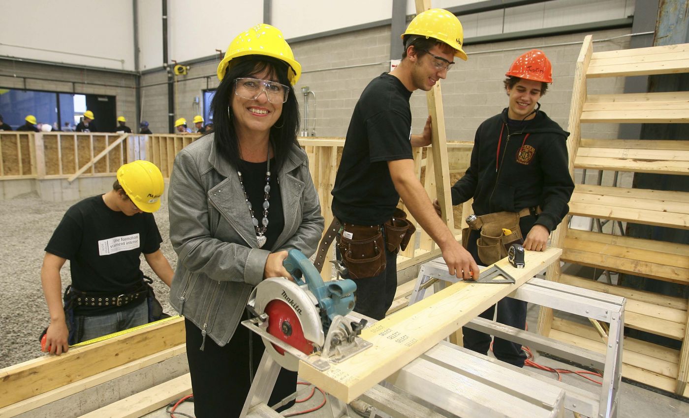 Colour photograph showing three young men and a middle-aged woman in a carpentry workshop, holding different tools. They are wearing hard hats, some also wearing protective glasses, and working around a wooden board. More workers are visible in the background.