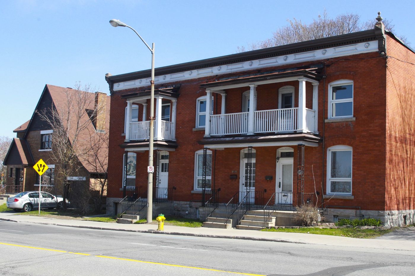 Photographie en couleur d’une maison en rangée à deux étages, à toit plat, en brique. La maison compte trois portes au rez-de-chaussée, surplombées de petits balcons. À sa gauche, une maison individuelle, à deux étages.
