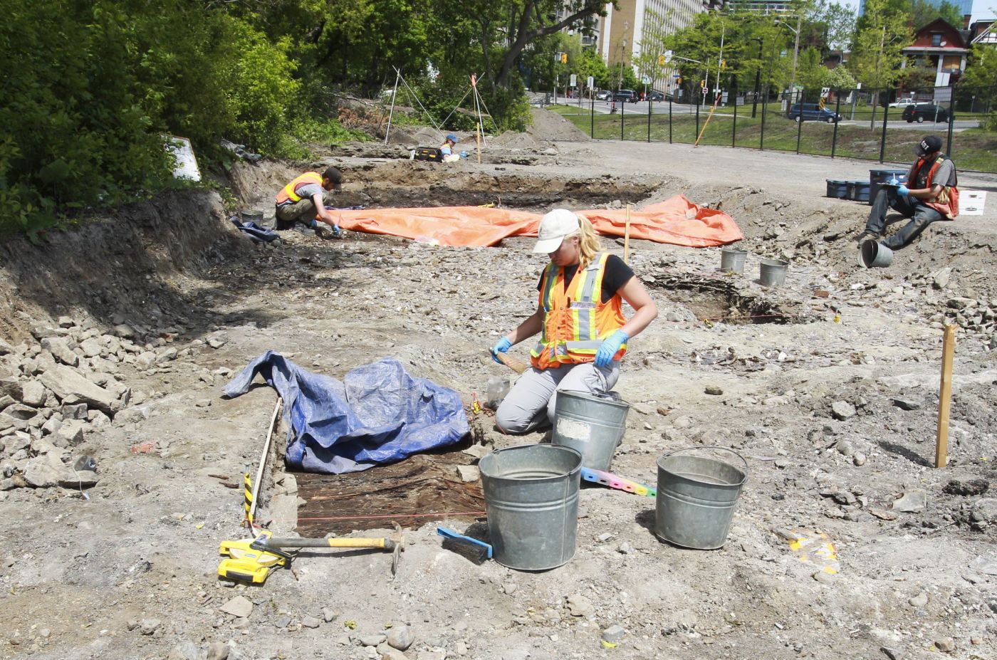 Colour photograph of a young woman kneeling, cleaning an artefact at an archaeological site. Two young men work behind her.