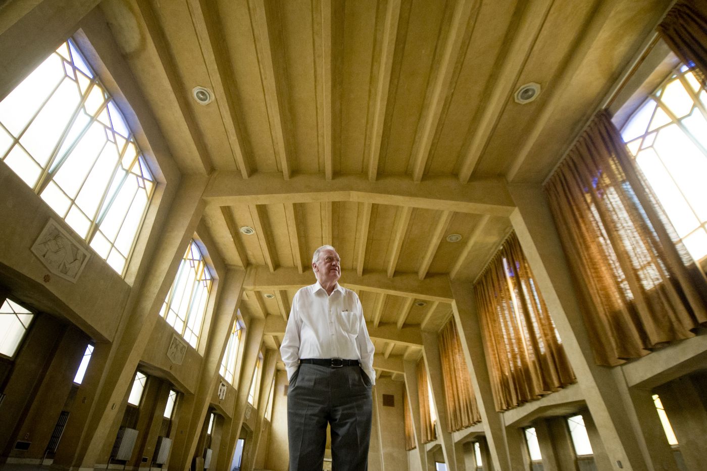 Colour photograph of an elderly man with white hair, standing in the center of a church, hands in the pockets of his pants. The building is simply decorated, with stained glass windows and a Way of the Cross.