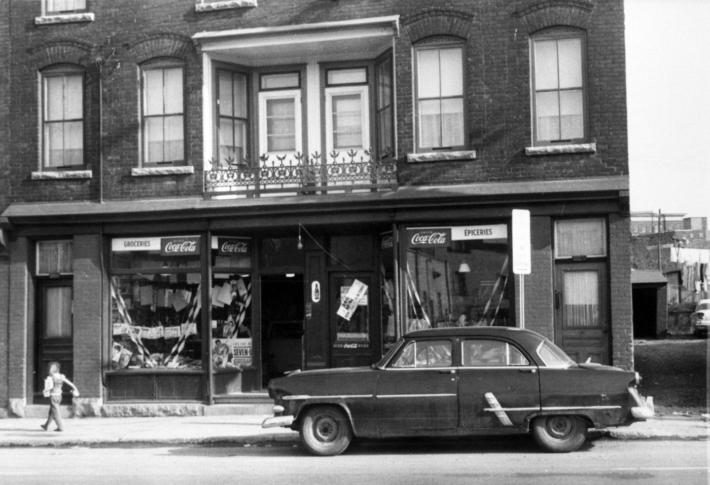 Photographie noir et blanc d’un jeune garçon marchant devant une maison en brique à plusieurs étages, abritant une épicerie au rez-de-chaussée. Les enseignes sont bilingues. Une voiture est stationnée devant le commerce.
